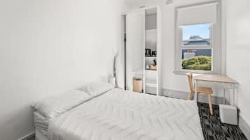 Un dormitorio con cama, escritorio y ventana que ofrece vista a un edificio y vegetación.