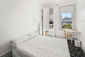 A bedroom with a bed, a desk, and a window with a view of a building and greenery.