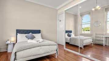 Un dormitorio con un espejo grande, piso de madera, una cama con ropa de cama blanca y una mesita de noche con lámpara.