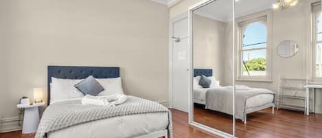 A bedroom with a large mirror, wooden flooring, a bed with white bedding, and a nightstand with a lamp.