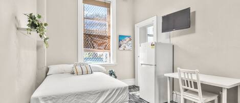 A bedroom with a bed, a desk, a chair, a refrigerator, a window with a view, and a television on the wall.