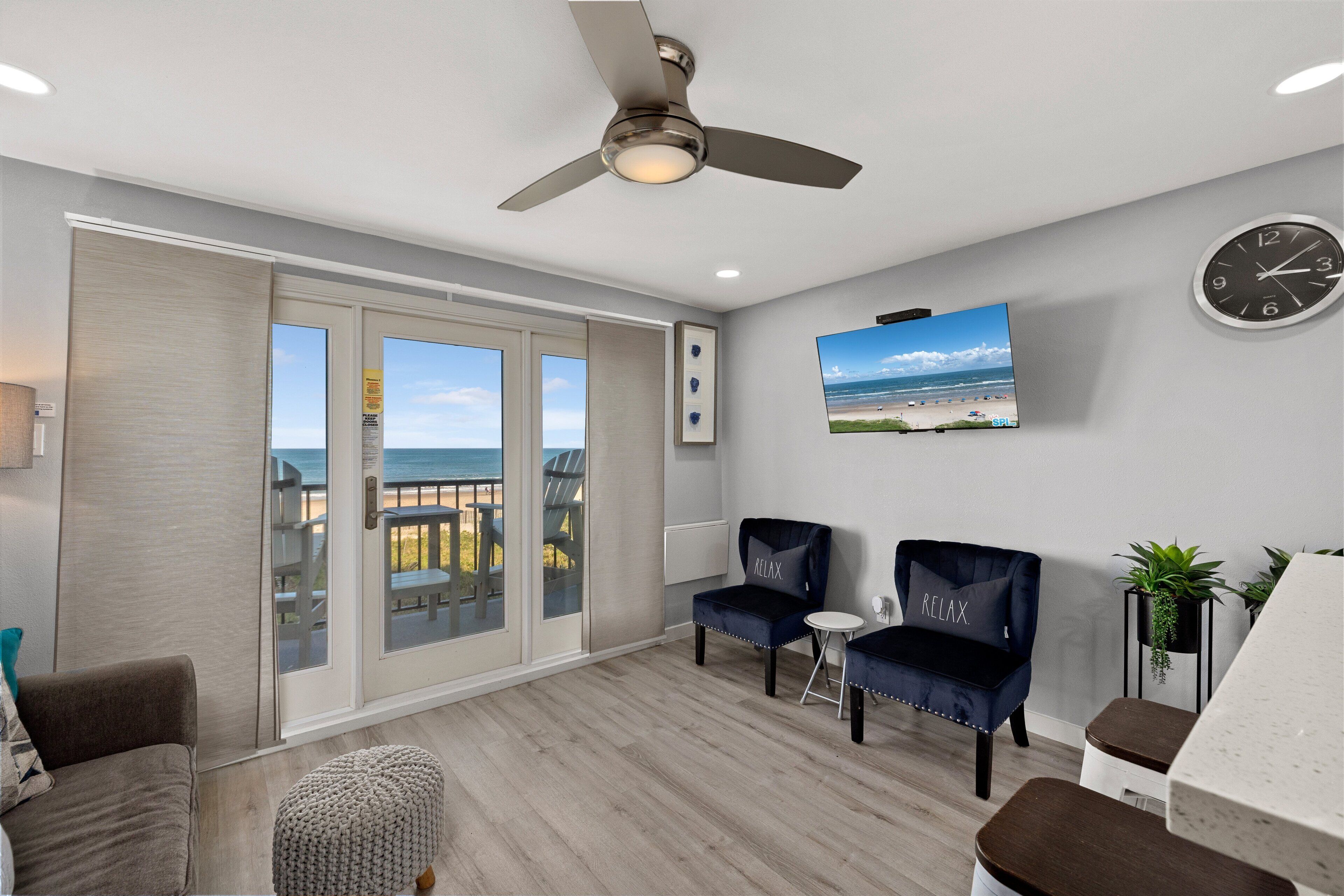 A spacious room with a balcony, a ceiling fan, a clock, and a TV displaying a beach scene.