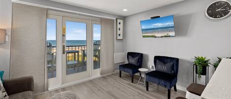A spacious room with a balcony, a ceiling fan, a clock, and a TV displaying a beach scene.
