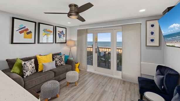 Ein modernes Wohnzimmer mit einem grauen Sofa, bunten Kissen, einem Deckenventilator und einem großen Fenster mit Blick auf den Strand.