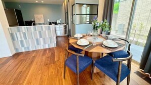 A modern dining area with a wooden table set for four, a kitchen in the background, and a large window providing a view of the outdoors.