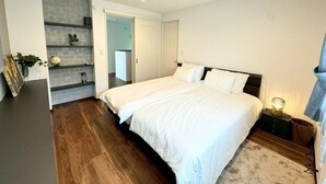 A bedroom with a large bed, a bedside table with a lamp, a small cabinet, and a door leading to another room.