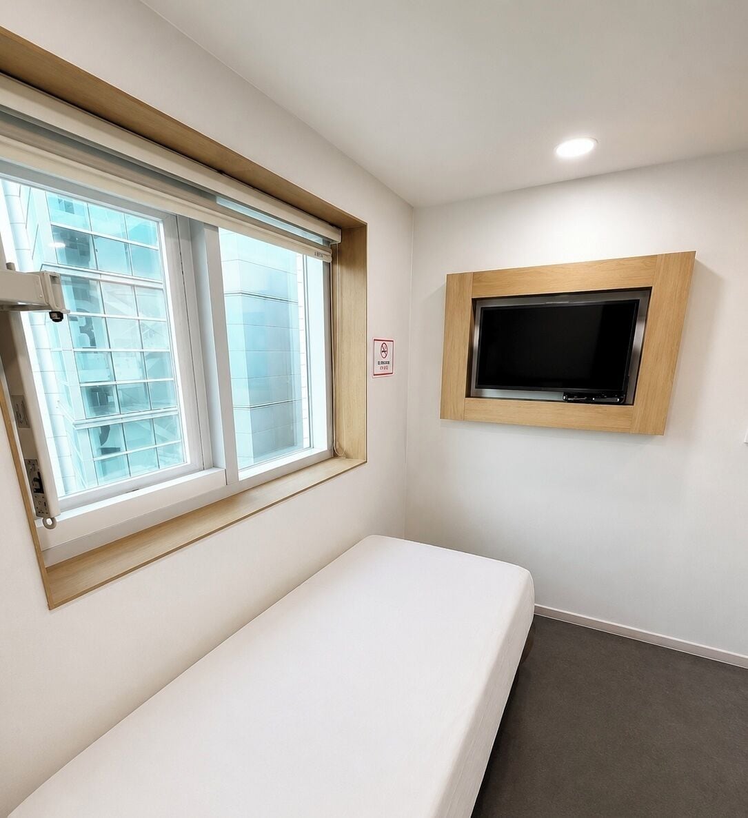 A small room with a single bed, a window with blinds, a mounted television, and a light switch on the wall.