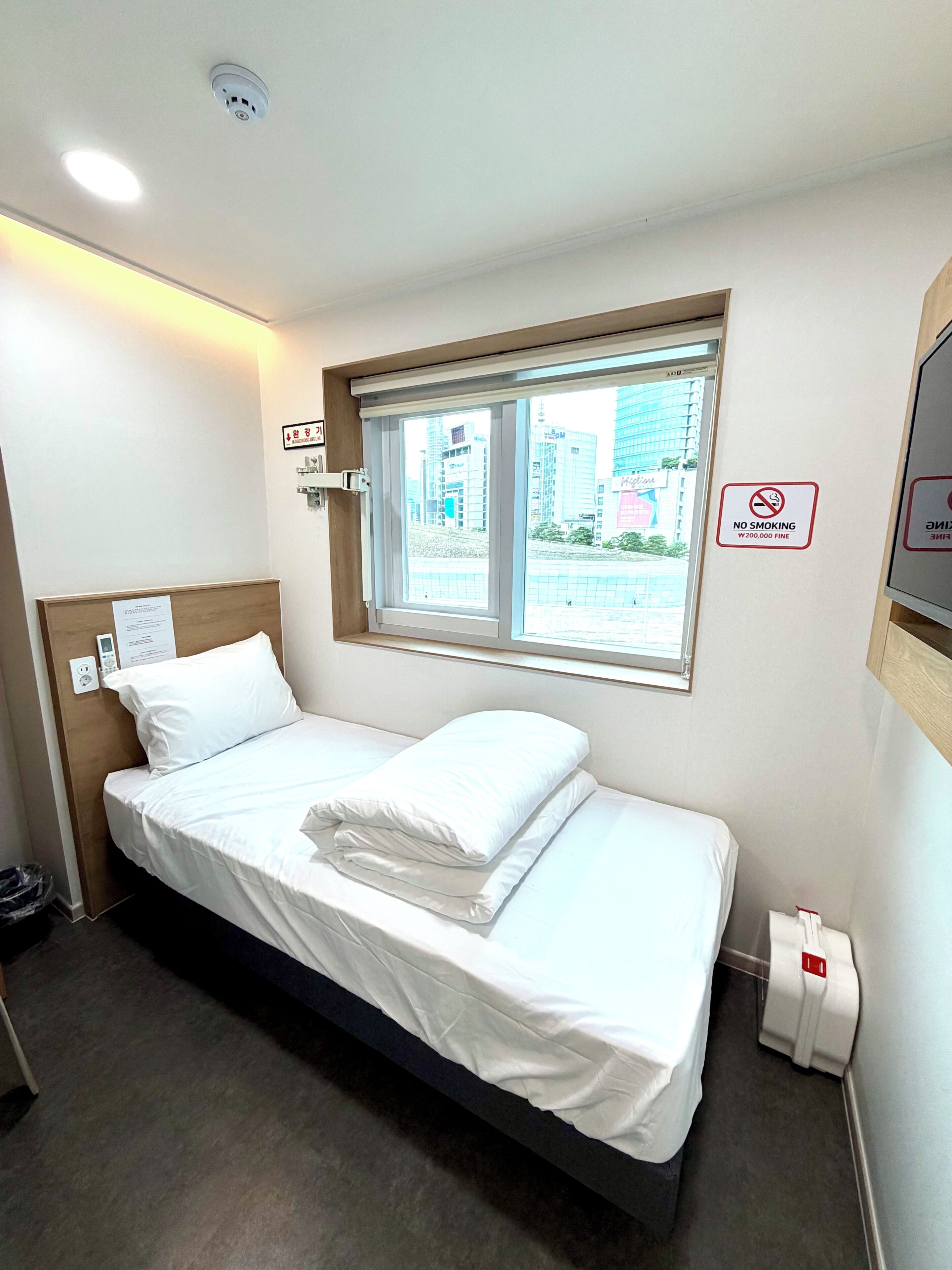 A hotel room with a bed, a window with a view of buildings, a 'No Smoking' sign, and a wall-mounted light fixture.