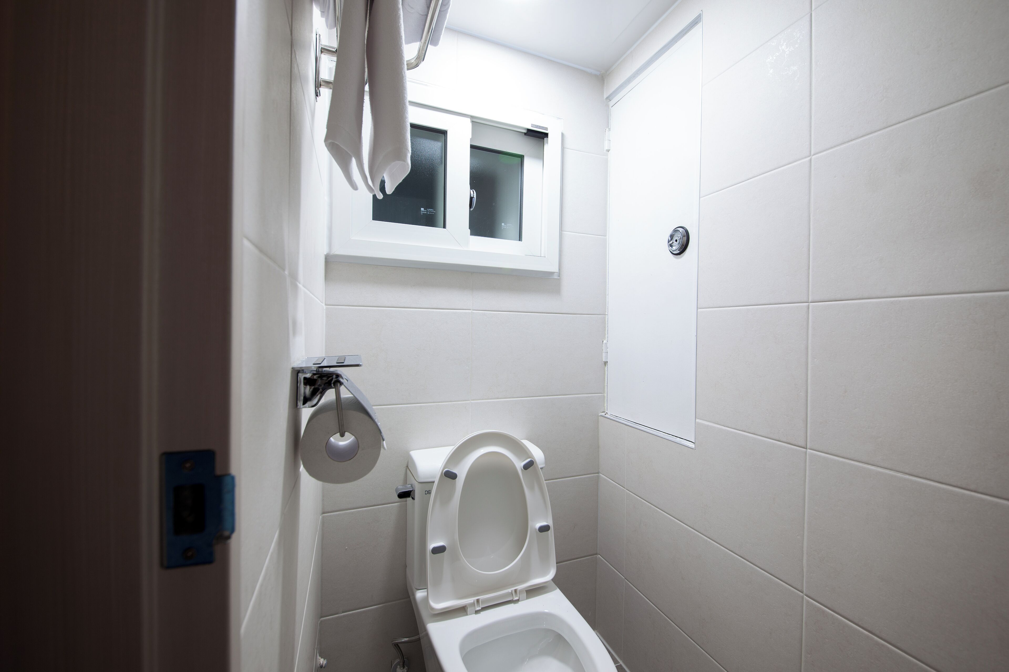 A small, clean bathroom with white tiles, a toilet, a window, and a towel rack.