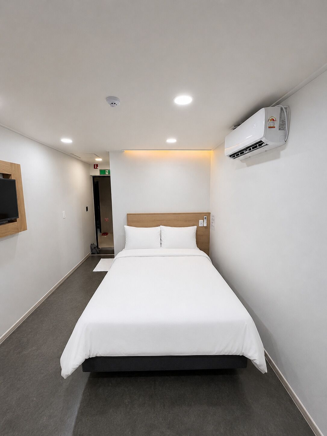 A hotel room with a bed, an air conditioning unit, a television mounted on the wall, and a door leading to another room.
