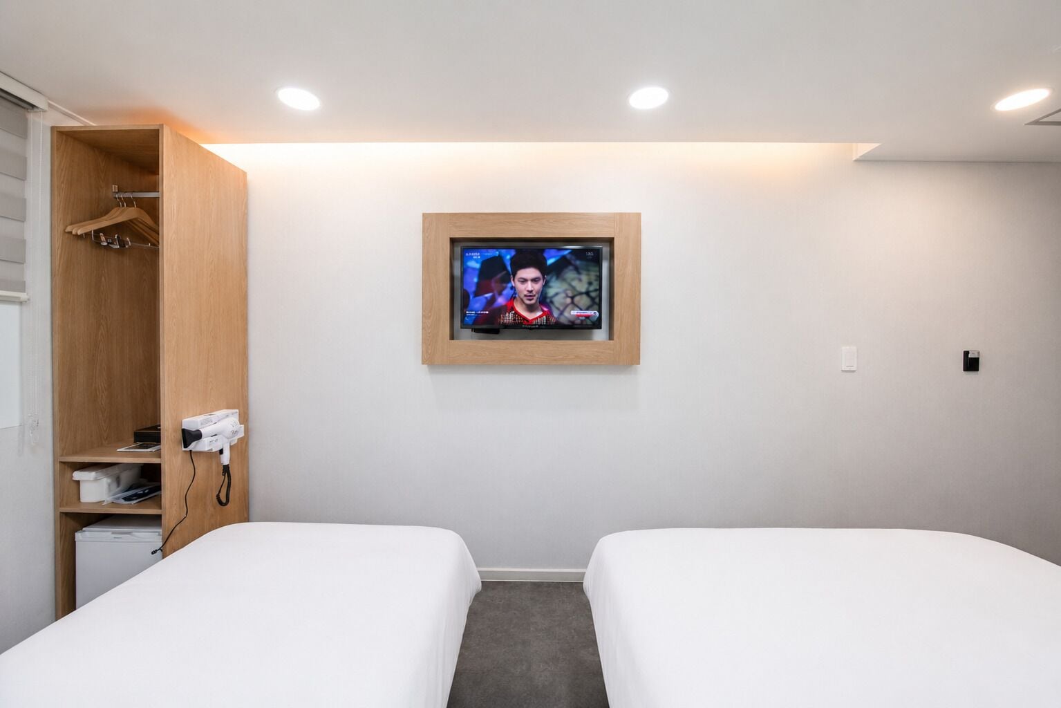 Quarto de hotel com duas camas, um guarda-roupa de madeira, uma televisão fixada na parede e um secador de cabelo ligado.