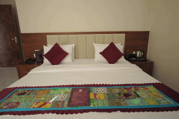 Habitación de hotel con una cama grande, dos almohadas, una mesita de noche con teléfono y cafetera, y un colcha decorativa en la cama.