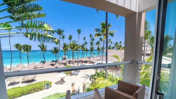 A balcony with wicker chairs overlooking a beach with palm trees and a clear blue sky.