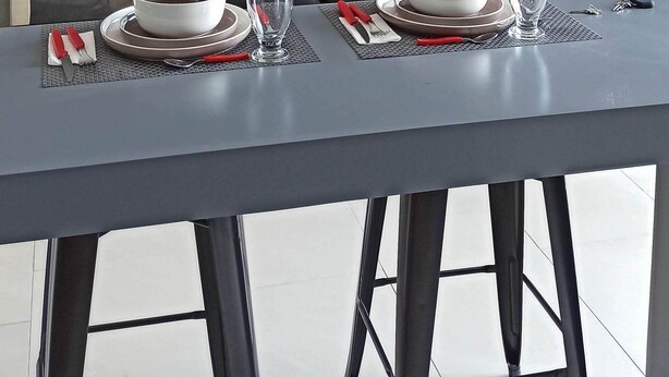 A modern living room with a dining area set for two. There is a sofa, a coffee table, and a dining table with plates, glasses, and cutlery. The room has large windows and a view of the outside.