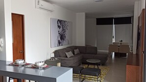 A modern living room with a grey sofa, a dining table with a bowl and cutlery, and a painting on the wall.