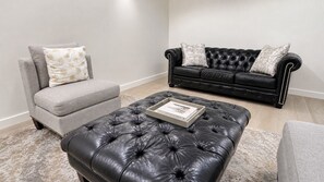 Un salon avec un fauteuil gris, un pouf en cuir noir et un canapé en cuir noir agrémenté de coussins à motifs.