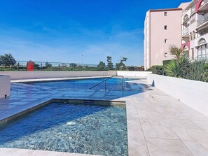 Una piscina con trampolino, circondata da un solarium piastrellato e un edificio con balconi.
