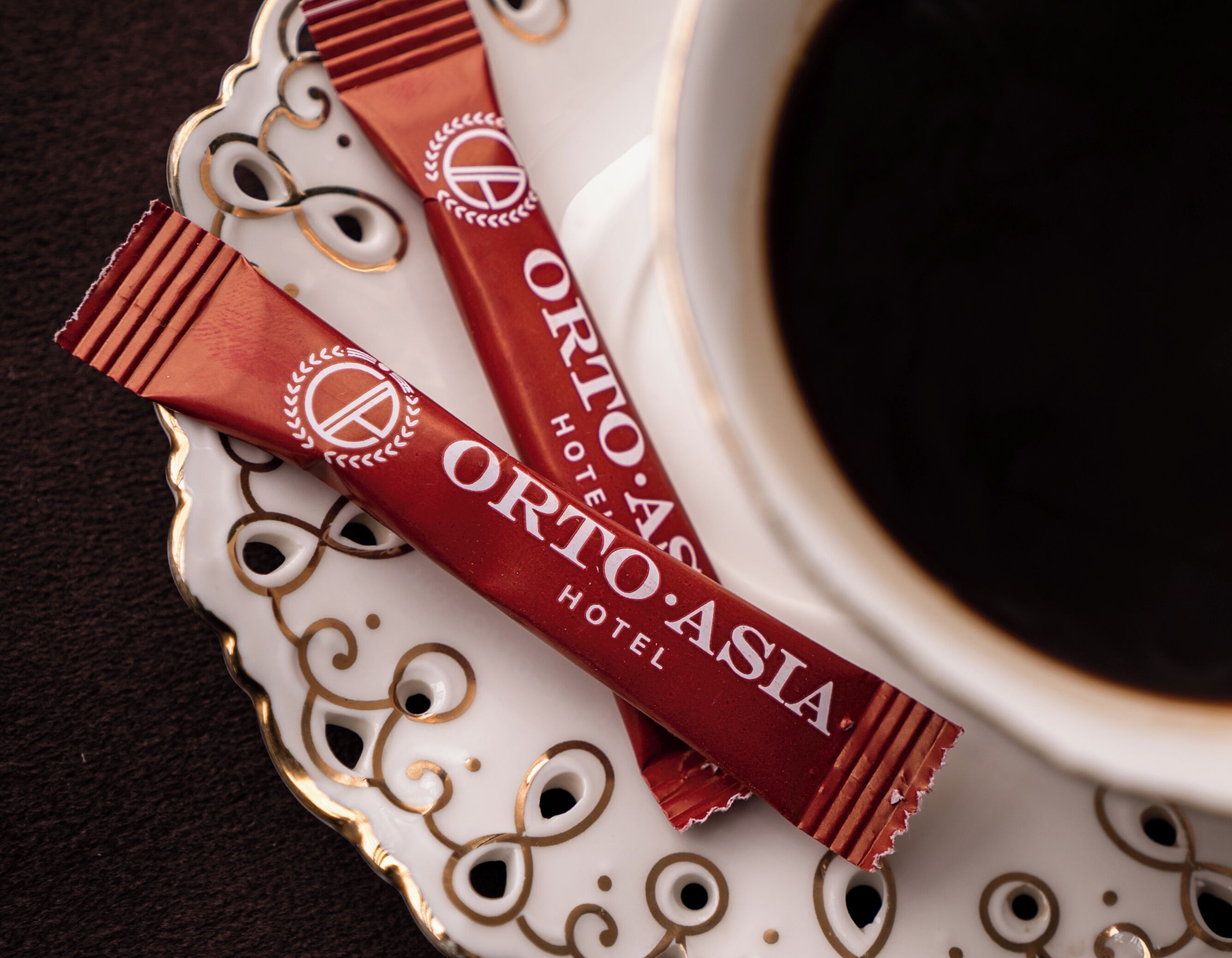 A white ceramic cup with a gold rim, containing a dark liquid, possibly coffee, and two chocolate bars labeled "ORTO AS HOTEL ORTOASIA".