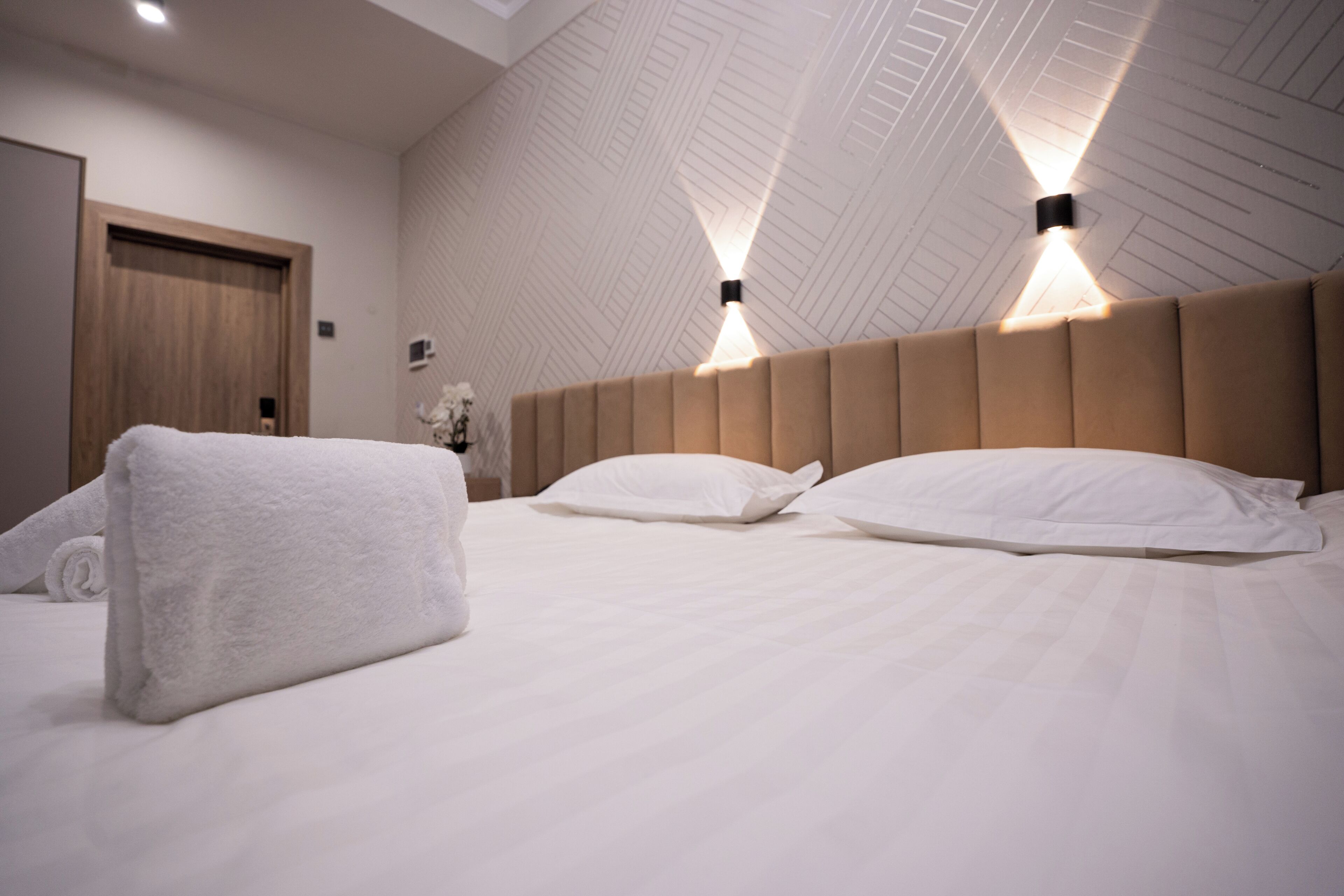 Quarto de hotel com uma cama grande, cabeceira de madeira e duas luzes fixadas na parede.