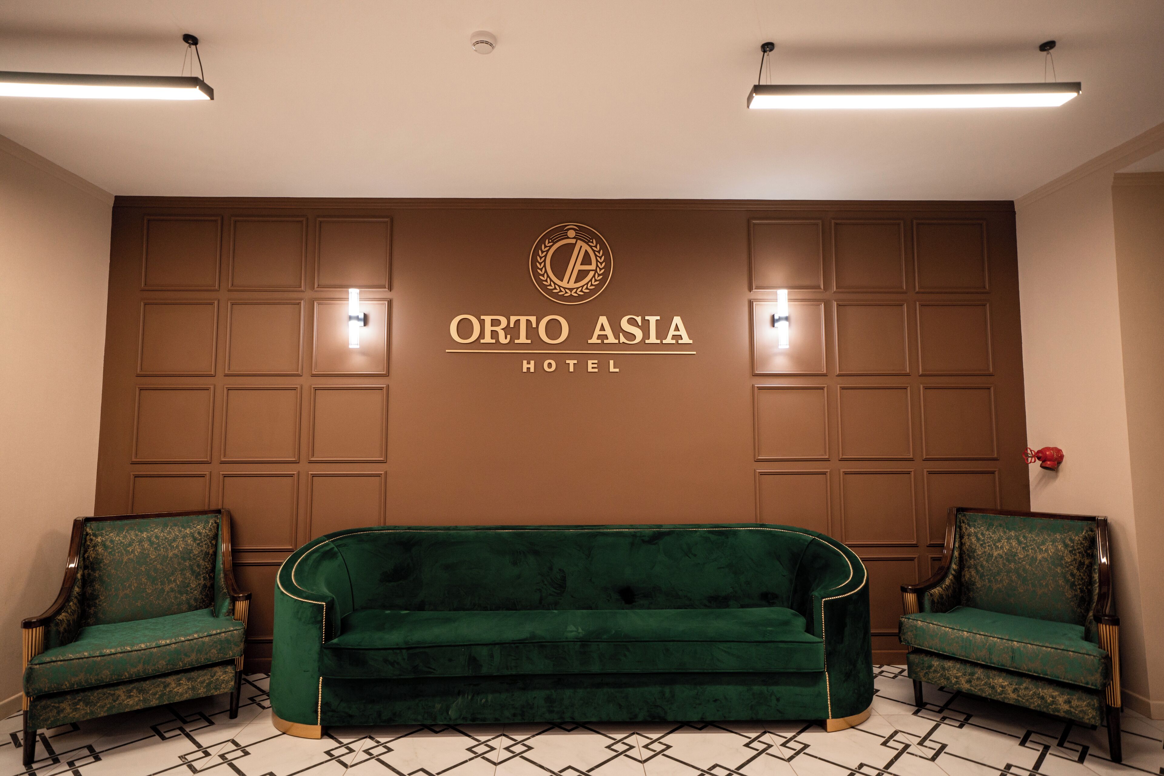 A hotel lobby with a green sofa, two green armchairs, and a wall-mounted sign reading "ORTO ASIA HOTEL".