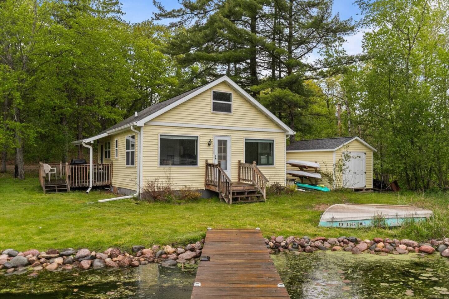 A single-story house with a front deck, a small shed, and a dock leading to a pond.