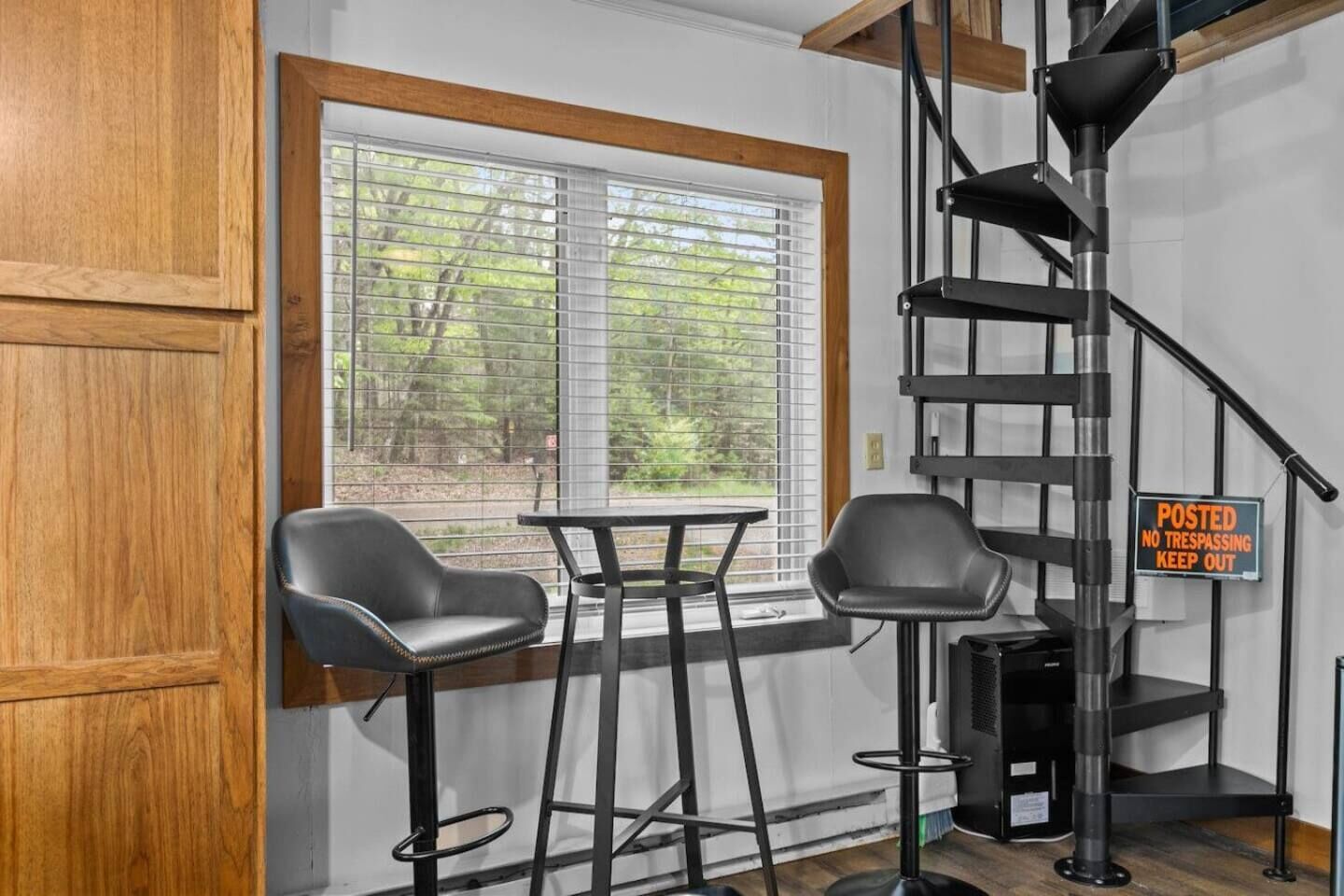 A room with a wooden door, a window with blinds, two black chairs, a small table, a black metal staircase, and a 'No Trespassing' sign.