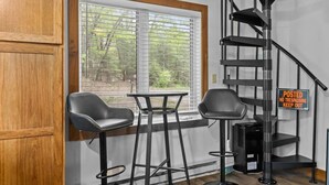 Ein Zimmer mit Holztür, einem Fenster mit Jalousien, zwei schwarzen Stühlen, einem kleinen Tisch, einer schwarzen Metalltreppe und einem 'No Trespassing'-Schild.