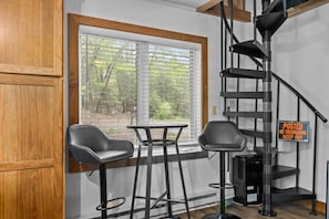 A room with a wooden door, a window with blinds, two black chairs, a small table, a black metal staircase, and a 'No Trespassing' sign.