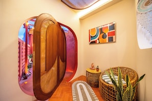 A hallway with a wooden acoustic panel, a colorful abstract painting, wicker furniture, and a patterned rug. - Kana Casa Canggu by Nagisa Bali (Canggu)