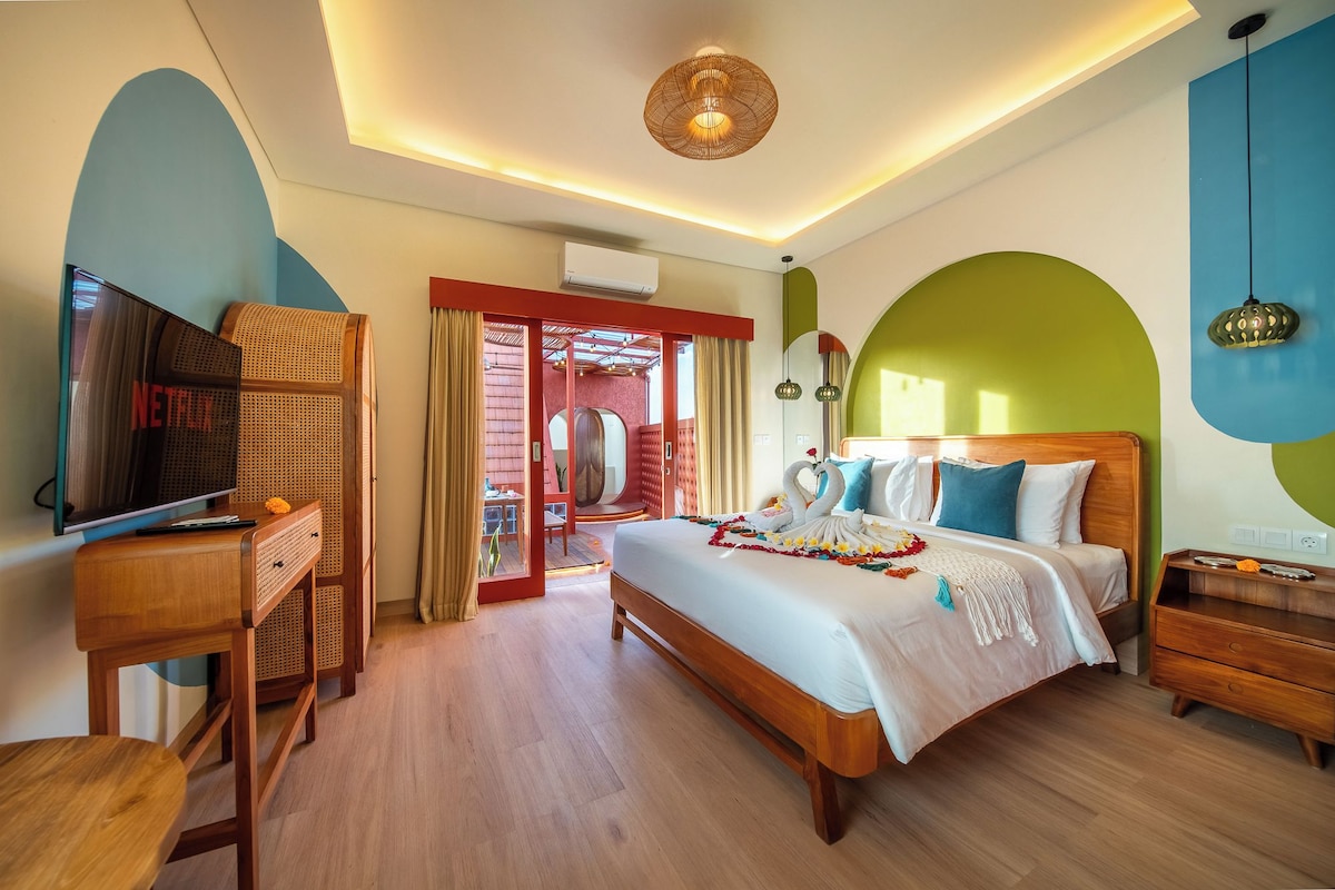 A hotel room with a bed, a TV, a wooden desk, and a door leading to an outdoor area.