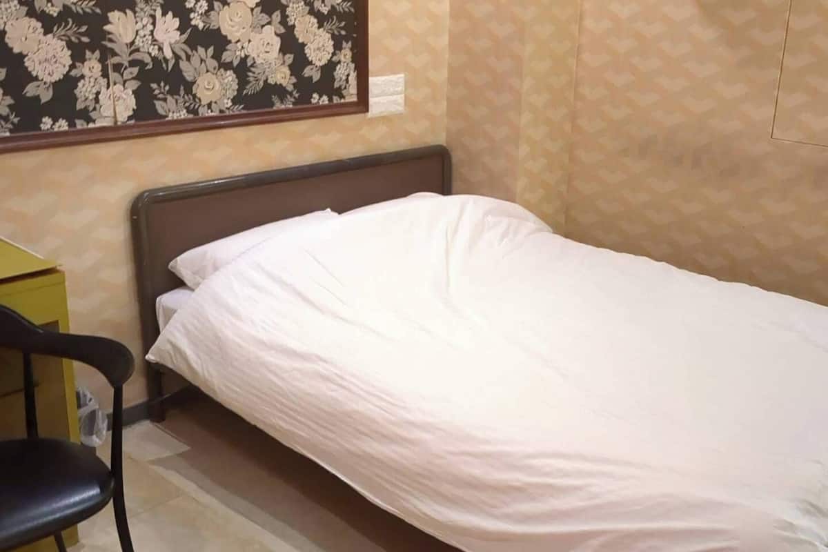 Un lit simple avec une literie blanche, dans une pièce dont les murs sont recouverts d’un papier peint à motifs et où se trouve un petit bureau.