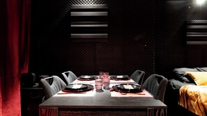 A dining area with a wooden table set for four, surrounded by chairs, in a room with a patterned wall and a bed with a yellow blanket. - Cinéma Provence - Air-conditioned - 7ᵉ art (Avignon)