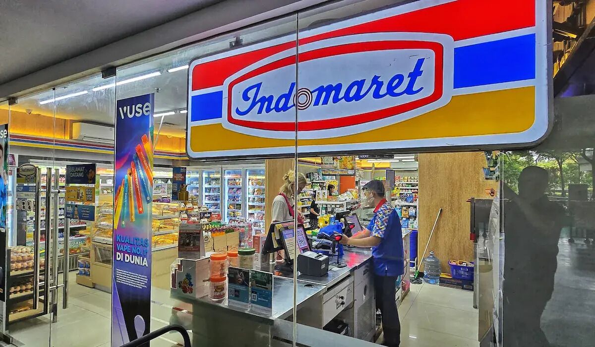 A supermarket interior with a prominent Indomaret sign, a variety of products on shelves, and a checkout counter.