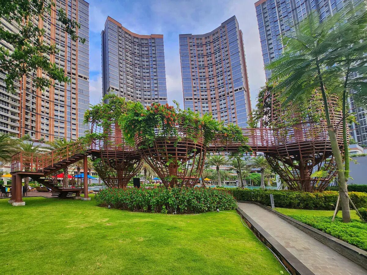 A modern urban park with a unique metal bridge structure amidst lush greenery, surrounded by high-rise buildings.