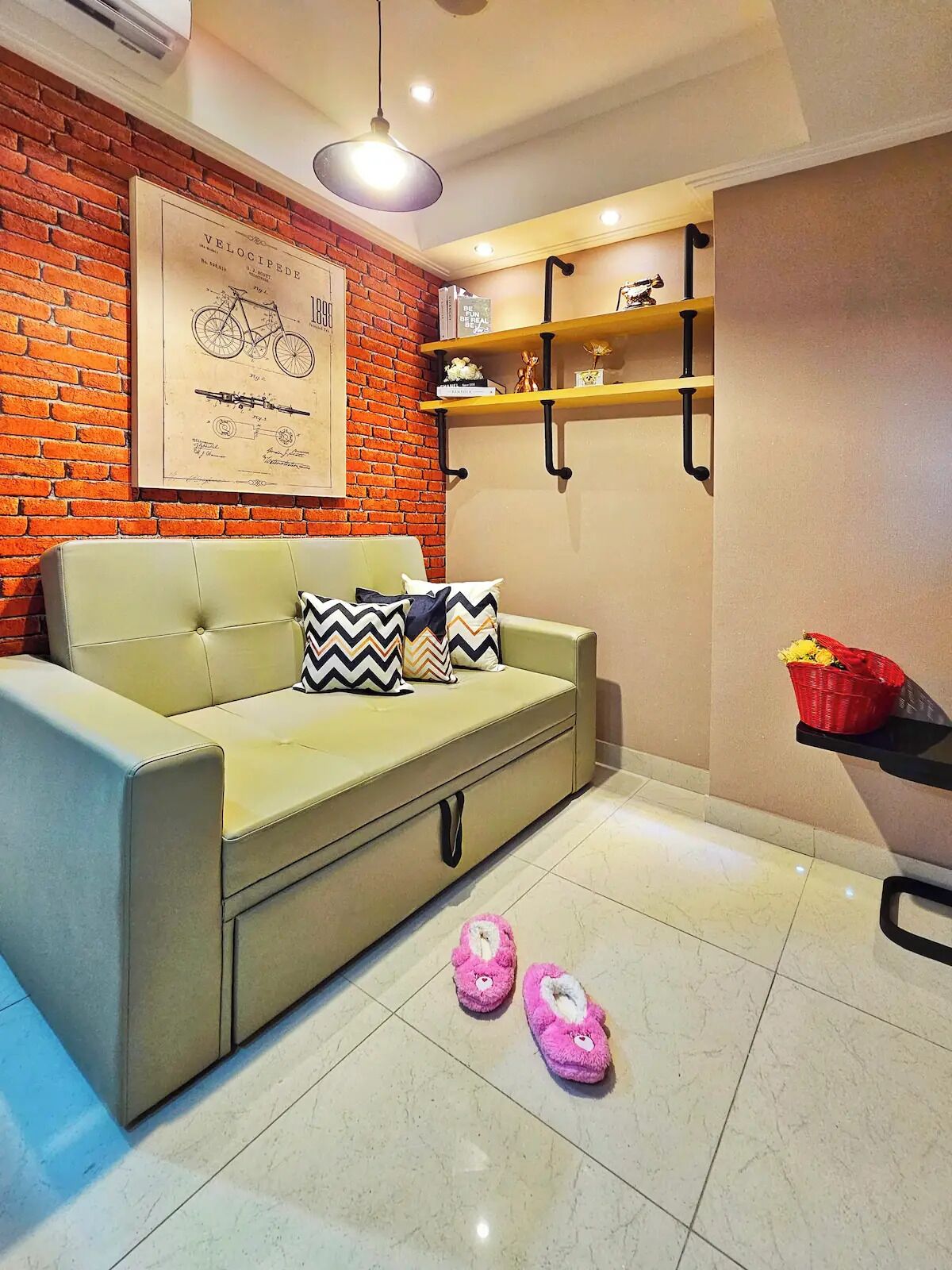 A living room with a brick wall, a sofa, a framed blueprint, shelves, and slippers on the floor.