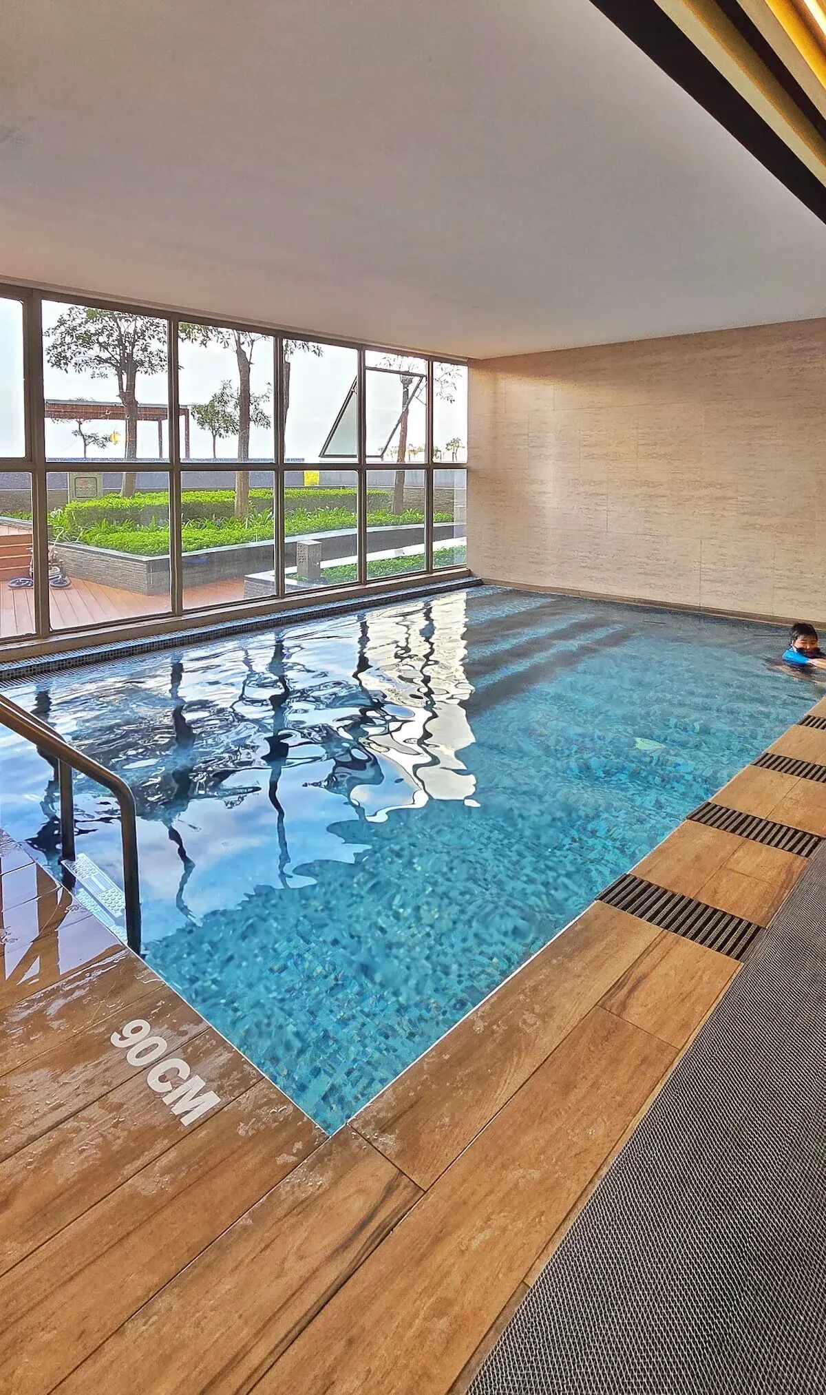 An indoor swimming pool with clear blue water, wooden deck, and large windows offering a view of greenery outside.
