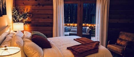 A cozy bedroom with a large bed, a nightstand with a lamp, a chair, and a view of the outdoors through a sliding door.