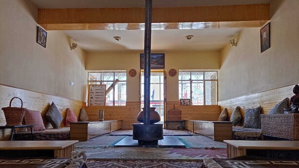 A traditional room with a central fireplace, wooden benches, and patterned rugs. - Juniper Home Kaza By Mountain Bee (Spiti)