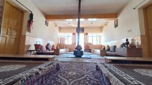 A spacious room with a high ceiling, a central pillar, and a patterned carpet. There are two sofas with cushions, a small table, and a wall-mounted shelf with decorative items. - Juniper Home Kaza By Mountain Bee (Spiti)