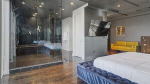 A modern bedroom with a large bed, a glass shower enclosure, and a flat-screen TV mounted on the wall.