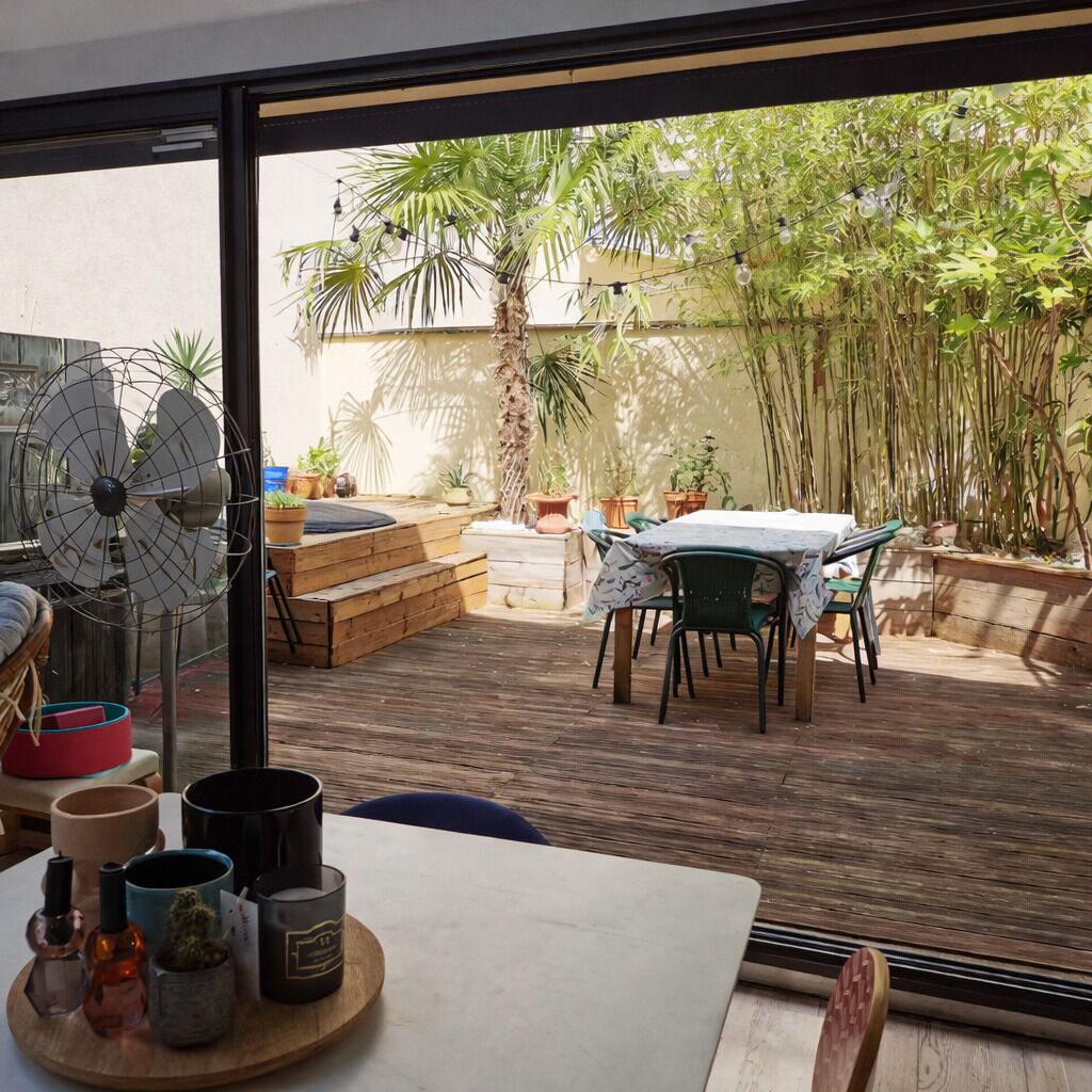 A room with a large window, wooden flooring, a fan, a table with candles, and a view of a garden with plants and a palm tree.