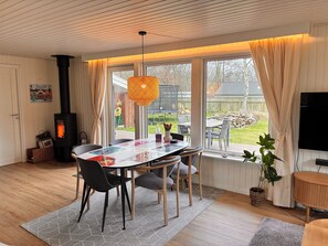 A cozy living room with a fireplace, a dining table with chairs, a TV, and a large window with curtains. - Lovely holiday home with garden near water (Rødby)
