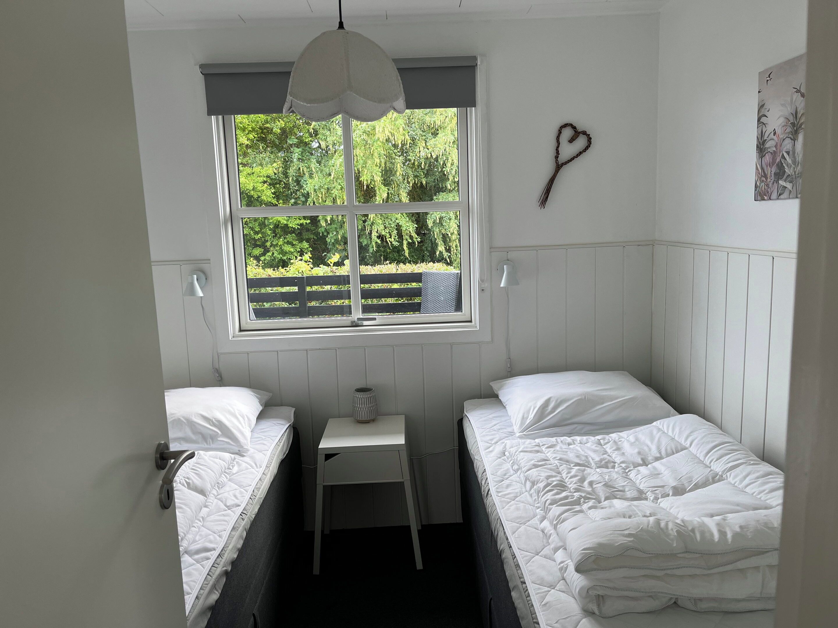 A room with two single beds, a small table, a window with a view of greenery, and a heart-shaped decoration on the wall.