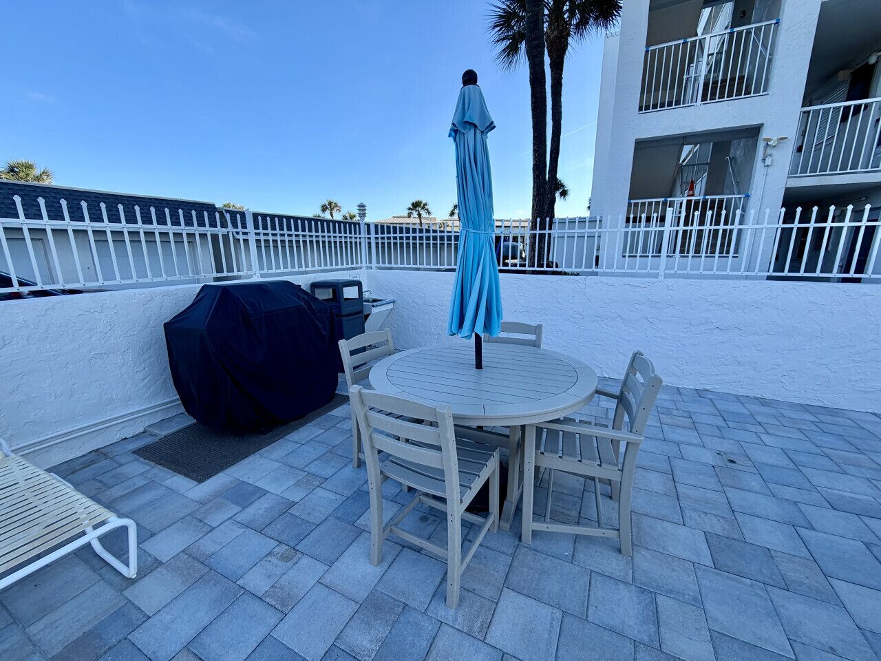 An outdoor patio with a round table and chairs, a covered grill, and a blue umbrella.
