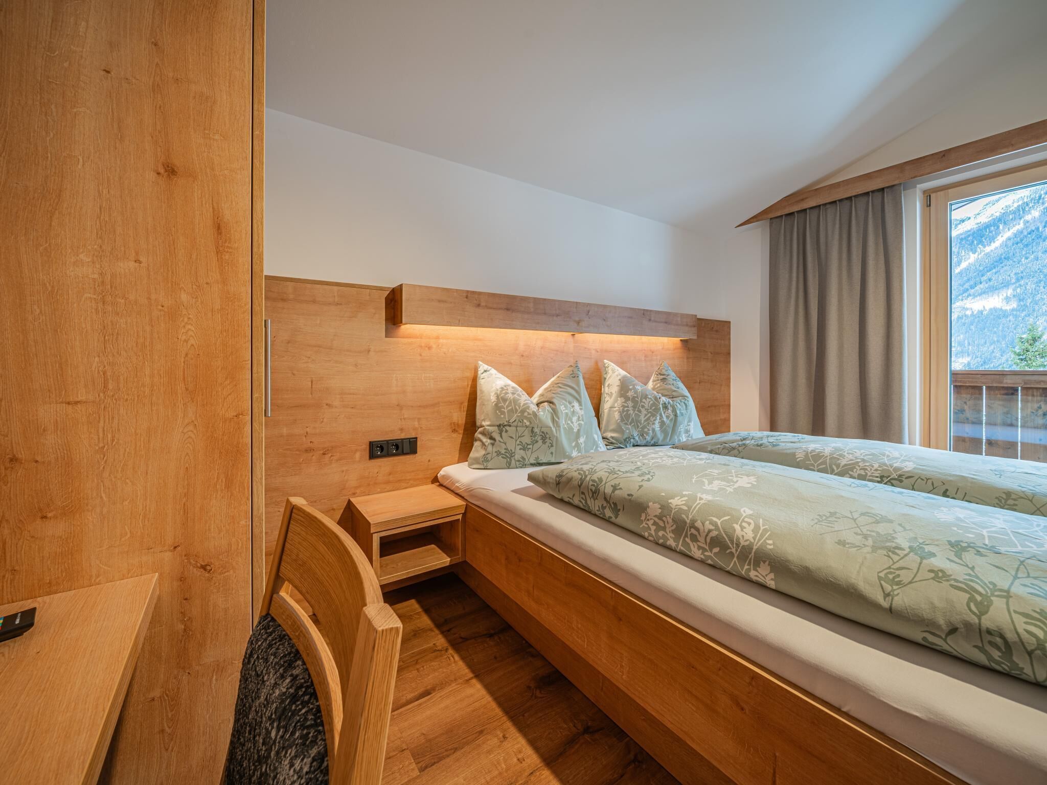 Une chambre moderne avec une tête de lit en bois, un lit recouvert d’une literie à motifs floraux, une table de chevet et une vue sur les montagnes par la fenêtre.