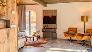 Uma sala de estar moderna com um sofá, duas poltronas, uma mesa de centro, uma TV fixada numa parede de madeira e uma janela ampla.