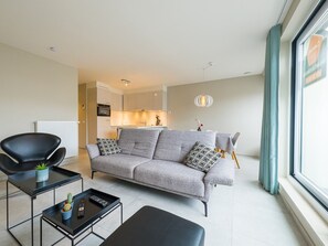 Ein modernes Wohnzimmer mit einem grauen Sofa, einem schwarzen Sessel und einem kleinen Couchtisch mit einem Kaktus. Im Hintergrund ist eine Küche zu sehen.