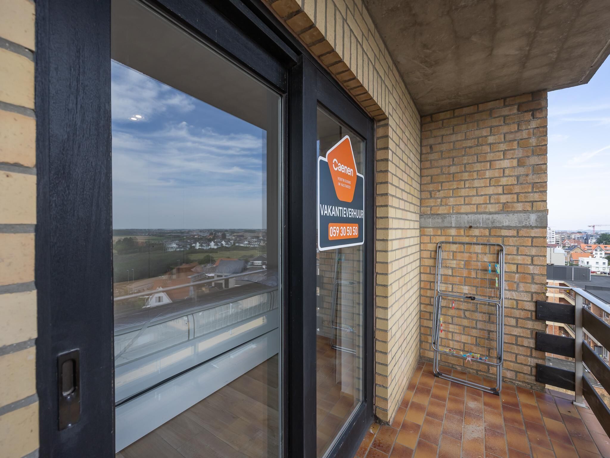 Blick von einem Balkon mit blauem Himmel, einer Ziegelwand und einem Schild für 'Caenen Vakantieverhuur' mit einer Telefonnummer.
