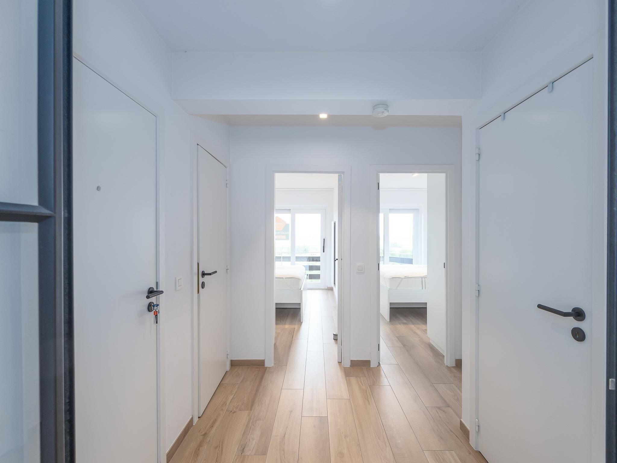 A hallway with white doors, a wooden floor, and a view into a room with a window.