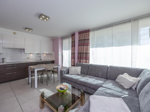 Ein modernes Wohnzimmer mit grauem Sofa, einem Glastisch und einer Küchenzeile mit weißen Schränken sowie einem Esstisch.